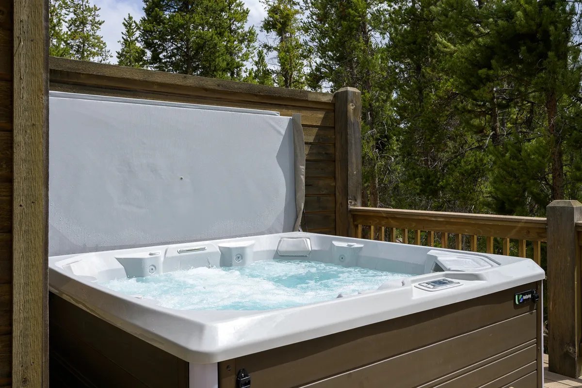 An inviting exterior view of a wooden deck surrounded by greenery, featuring a private hot tub and comfortable seating. The tranquil setting is perfect for relaxation, with scenic views and an atmosphere ideal for unwinding after a day of exploration.