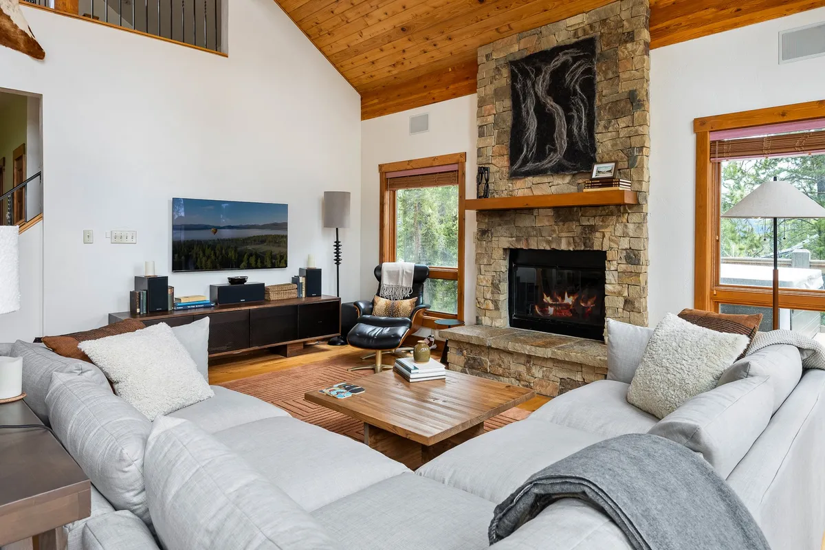 A spacious, light-filled living room featuring a large stone fireplace as its centerpiece. The comfortable sectional sofa and wooden coffee table are surrounded by expansive windows that frame serene forest views, creating a cozy and inviting atmosphere for relaxing, entertaining, or enjoying a movie night.