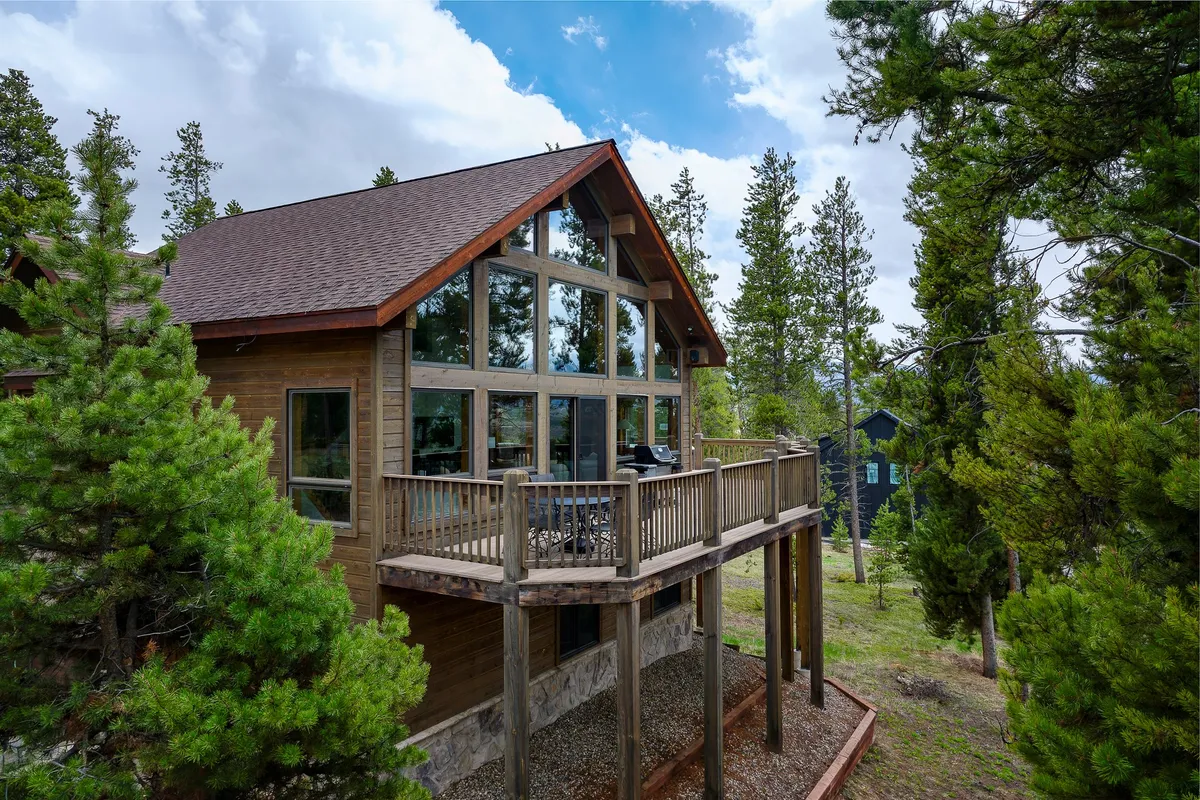 An exterior angle showcasing the wrap-around deck, large glass windows, and wooden exterior. The deck area is set up for outdoor relaxation, dining, and enjoying the surrounding natural beauty, with glimpses of sunset hues and native greenery.