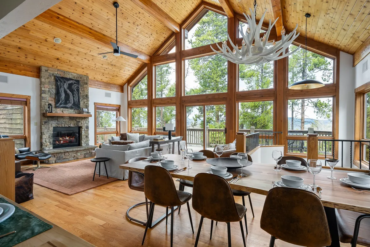 The image showcases the inviting interior of the Silver Lining Lodge, featuring a spacious living area with soaring windows revealing breathtaking mountain views, a striking stone fireplace, and a welcoming dining table ideal for family gatherings. Warm sunlight pours in, illuminating the cozy space and highlighting the perfect blend of rustic charm and modern comfort.