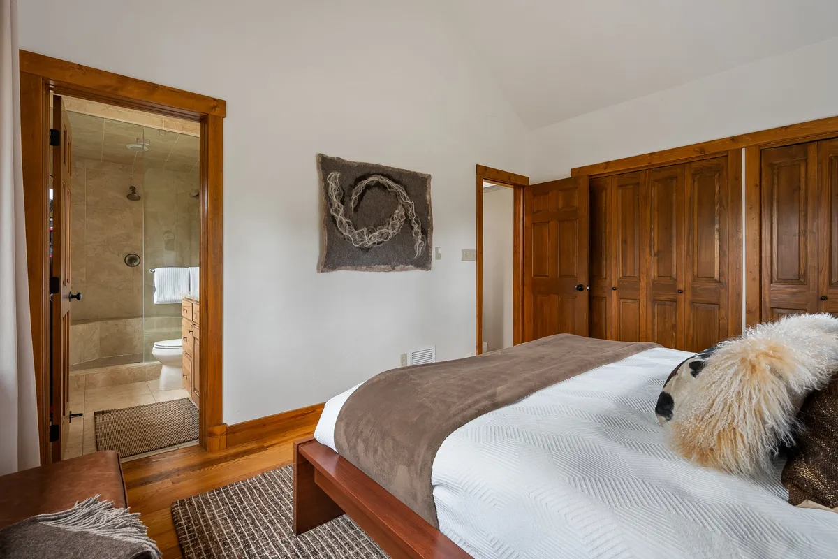 A bedroom with a distinctive wooden bed frame, textured wall art, plush pillows, and a view into an adjoining en-suite bathroom. The space feels warm and comfortable, with stylish wardrobe doors and soft lighting enriching the restful atmosphere.