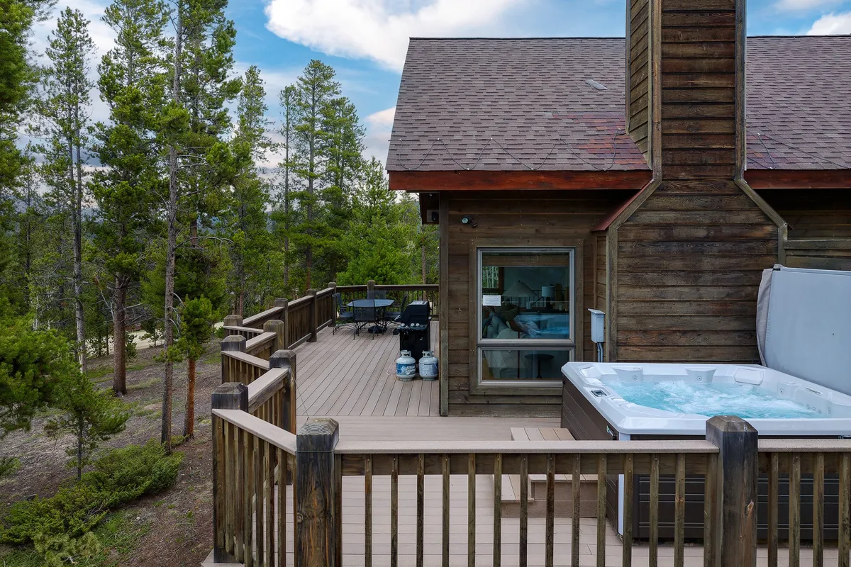An exterior view of a spacious wooden deck surrounded by trees, featuring a private hot tub overlooking the natural scenery. The area is inviting for relaxation, with the hot tub positioned for sunset views and stargazing.