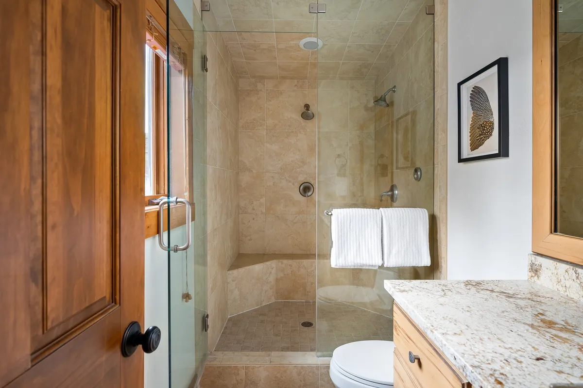 A luxurious bathroom featuring a glass shower, granite countertop, neatly stacked towels, and a wooden door. The tranquil setting evokes a spa-like atmosphere, ideal for refreshing and relaxing after a day of mountain adventure.