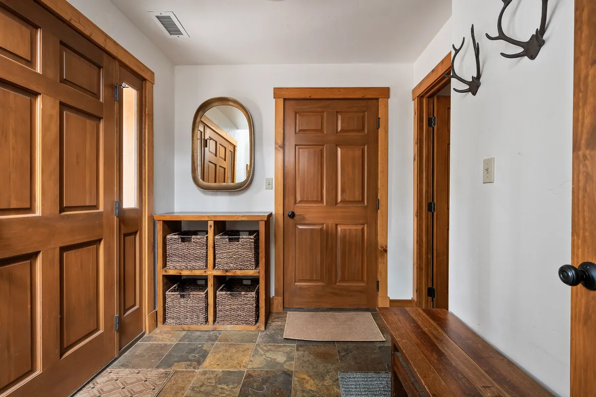 A welcoming and stylish entry area with wooden doors, decorative mirror, woven baskets, and antler decor on slate flooring. It’s designed as a functional yet inviting space where guests are greeted and can store outdoor gear.